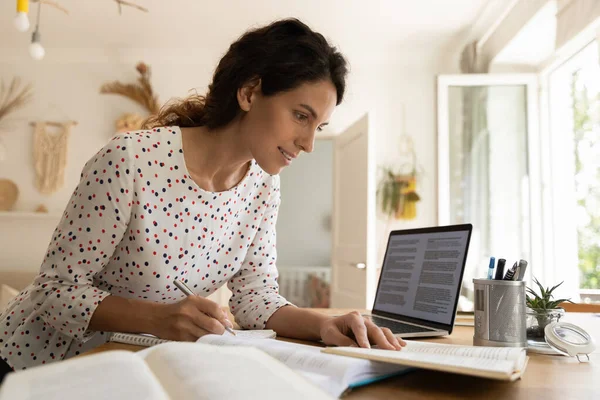 Улыбающаяся женщина, делающая заметки, пользующаяся ноутбуком, изучающая онлайн — стоковое фото