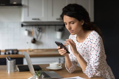 Akıllı telefon kullanan kendine güvenen bir kadın, masada dizüstü bilgisayarla oturuyor.