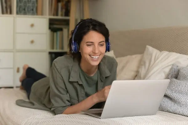 Mulher excitada em fones de ouvido sem fio usando laptop, deitada no sofá — Fotografia de Stock