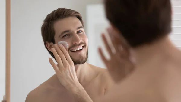 Happy handsome metrosexual guy applying sunscreen on face at mirror