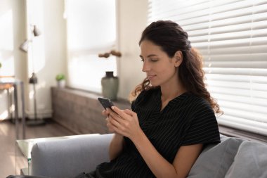 Genç kadın, modern akıllı telefon cihazıyla kanepeye oturur.