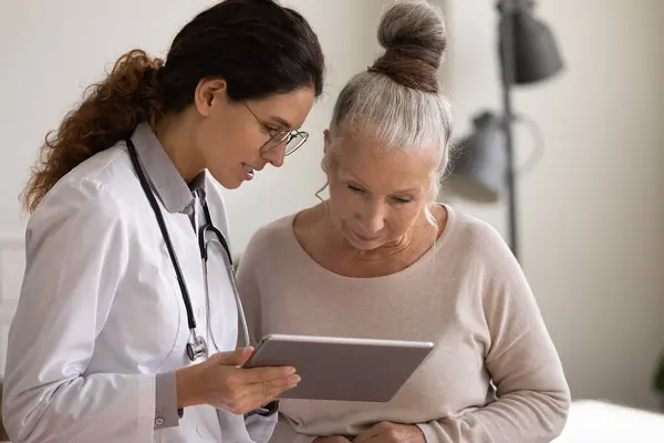 Vrouwelijke arts en oude patiënt gebruik pad gadget in kliniek — Stockfoto