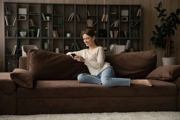Femme millénaire souriante jouant jeu mobile, reposant sur le canapé. — Photo