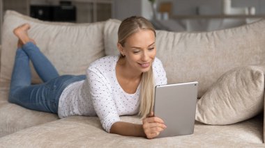 Tablet kullanan, rahat koltukta uzanan gülümseyen genç kadın.