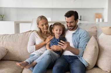 Küçük kızı olan mutlu bir aile, akıllı telefon kullanıyor ve koltukta oturuyor.