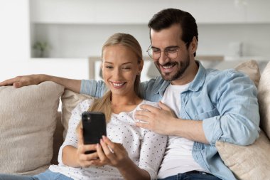 Gülümseyen genç bir çift akıllı telefon kullanarak sarılıyorlar koltukta birlikte oturuyorlar.