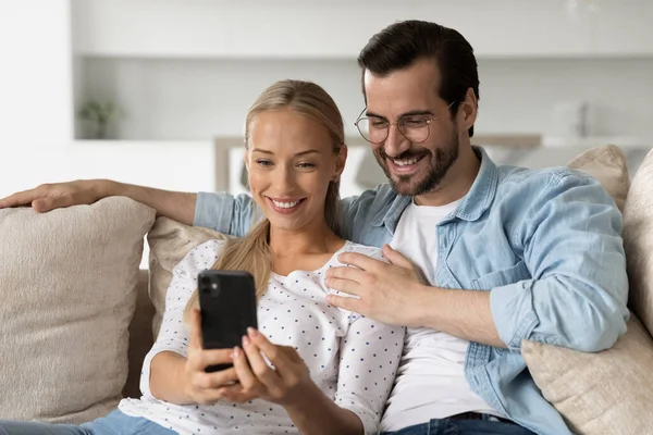Lächelndes junges Paar, das sich per Smartphone umarmt und gemeinsam auf der Couch sitzt — Stockfoto
