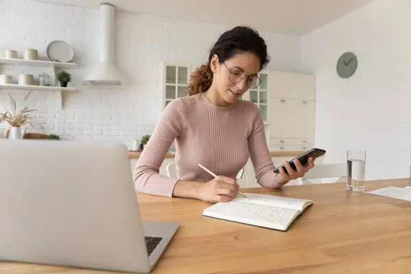 Självsäker ung kvinna skriva anteckningar, med hjälp av smartphone hemma. — Stockfoto