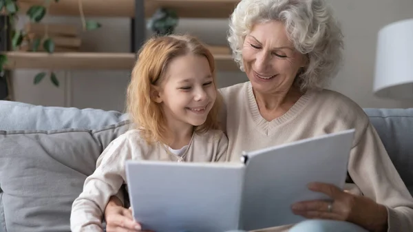 Sonriente nieta pequeña y madura abuela disfrutar interesante libro juntos —  Fotos de Stock
