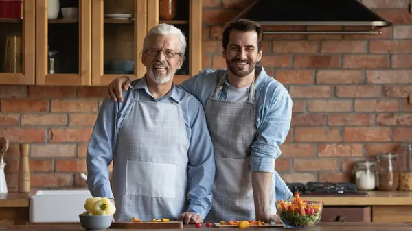 Feliz filho adulto e pai sênior em aventais cozinhar almoço — Fotografia de Stock