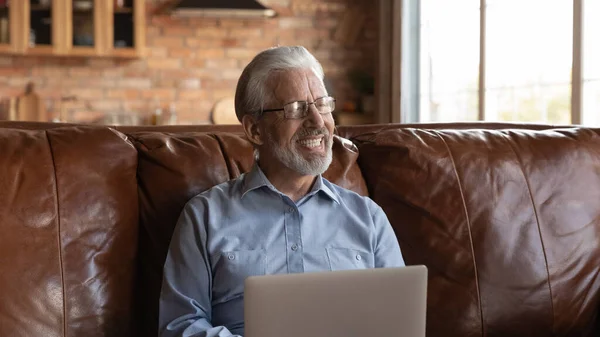 Счастливый пожилой 70-летний мужчина с ноутбуком, сидящий на диване — стоковое фото