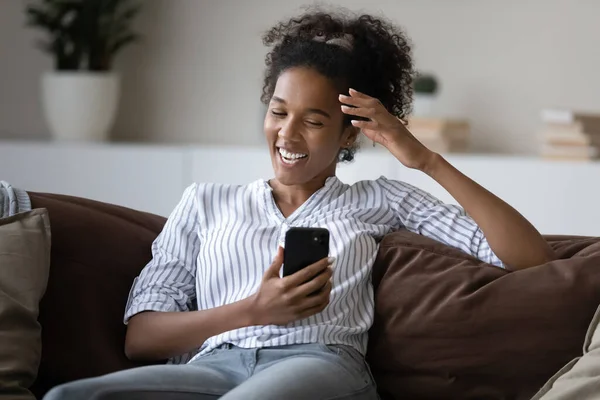 Excited African American woman using smartphone, sitting on cozy couch