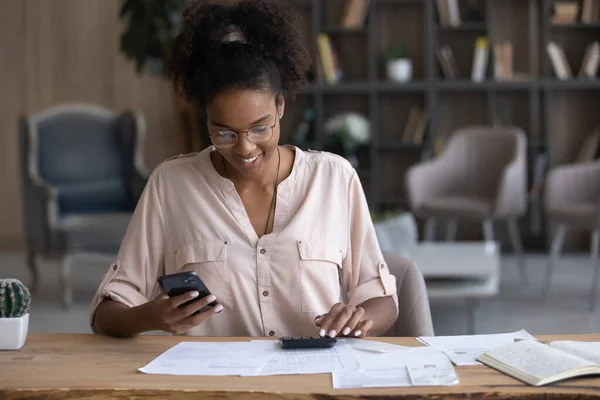 Femme afro-américaine souriante dans des lunettes de calcul des factures, en utilisant un smartphone — Photo