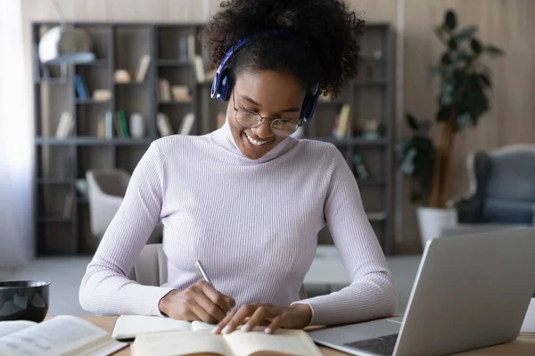 Smiling African American woman in headphones taking notes, using laptop — Stock Photo, Image
