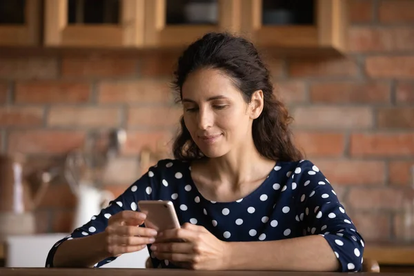 Focused Latin woman reading text on cellphone screen in kitchen — стоковое фото