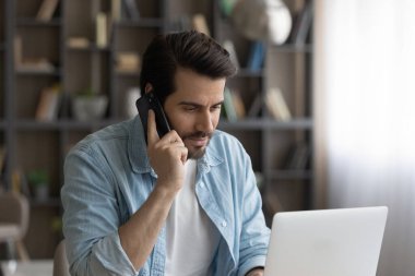 Konsantre genç adam telefonla konuşuyor, bilgisayarla çalışıyor..