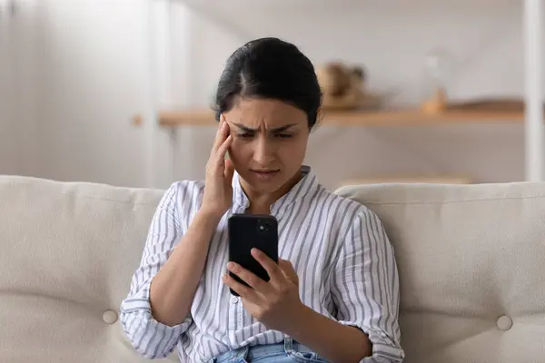 Unhappy Indian woman looking at phone screen, sitting on couch — Fotografia de Stock