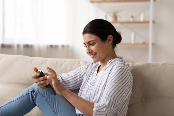 Smiling Indian millennial woman using smartphone, relaxing on couch — Stock fotografie
