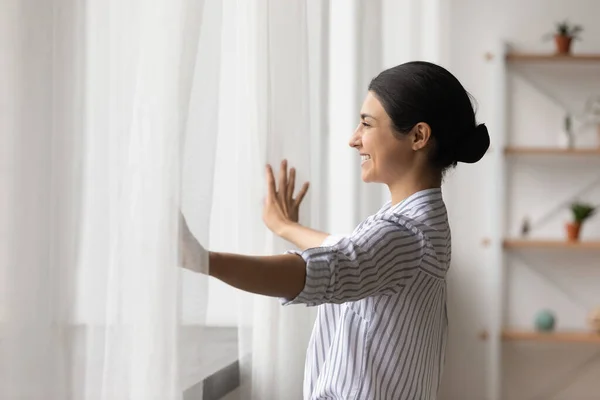 Smiling dreamy Indian woman opening curtains in early morning — Fotografia de Stock