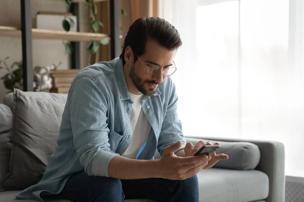 Man in glasses sit on sofa holding smartphone read messages