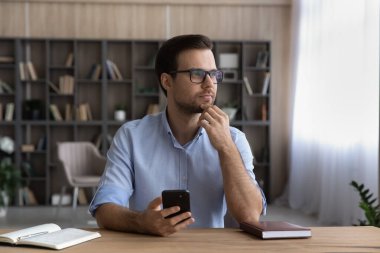 Hayalperest genç adam, masaya otur cep telefonunu tut ve yana bak.