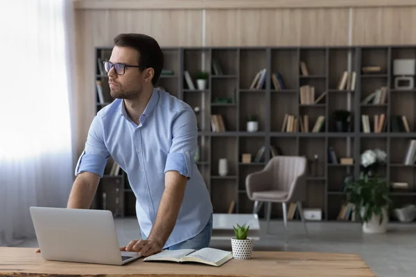 Hombre estudiante stand by desk con portátil mirar hacia otro lado pensar — Foto de Stock