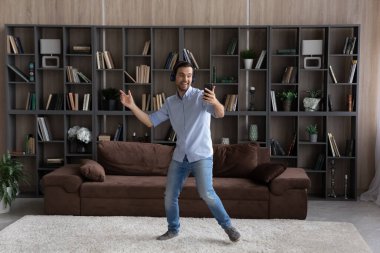 Kulaklıklı genç adam oturma odasında akıllı telefonuyla dans ediyor.