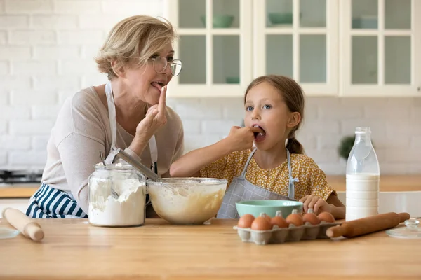 Menina curiosa com avó madura degustação de massa, cozinhar juntos — Fotografia de Stock