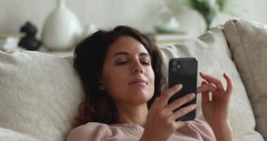 Gülümseyen, rahat, genç bir kadın evde akıllı telefon kullanıyor..