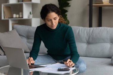 Laptop ve hesap makinesi kullanan odaklanmış bir kadın, yurt içi faturaları kontrol ediyor