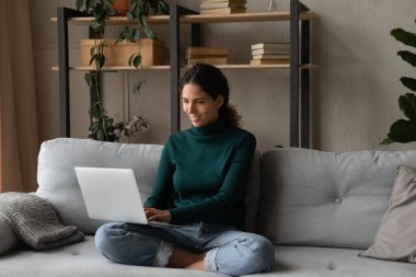 Kucağında dizüstü bilgisayarla kanepede dinlenen gülümseyen genç kadın.