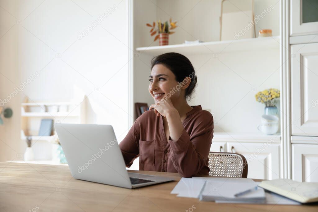 Smiling Indian woman work on laptop dreaming planning