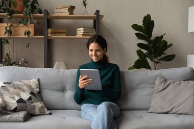 Gülümseyen kadın tablet tutuyor, uygulamalara bakıyor, daktilo yazıyor, kanepede oturuyor.