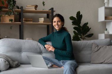 Portre gülümseyen beyaz kadın dizüstü bilgisayarla koltukta dinleniyor.