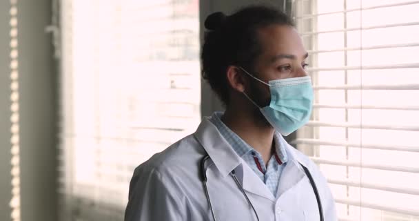 Pensive African male doctor wear uniform facemask looking out window — Stock Video