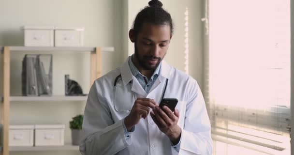 Усміхаючись, африканський лікар чоловічої статі ламається на роботі, користуючись мобільним телефоном. — стокове відео