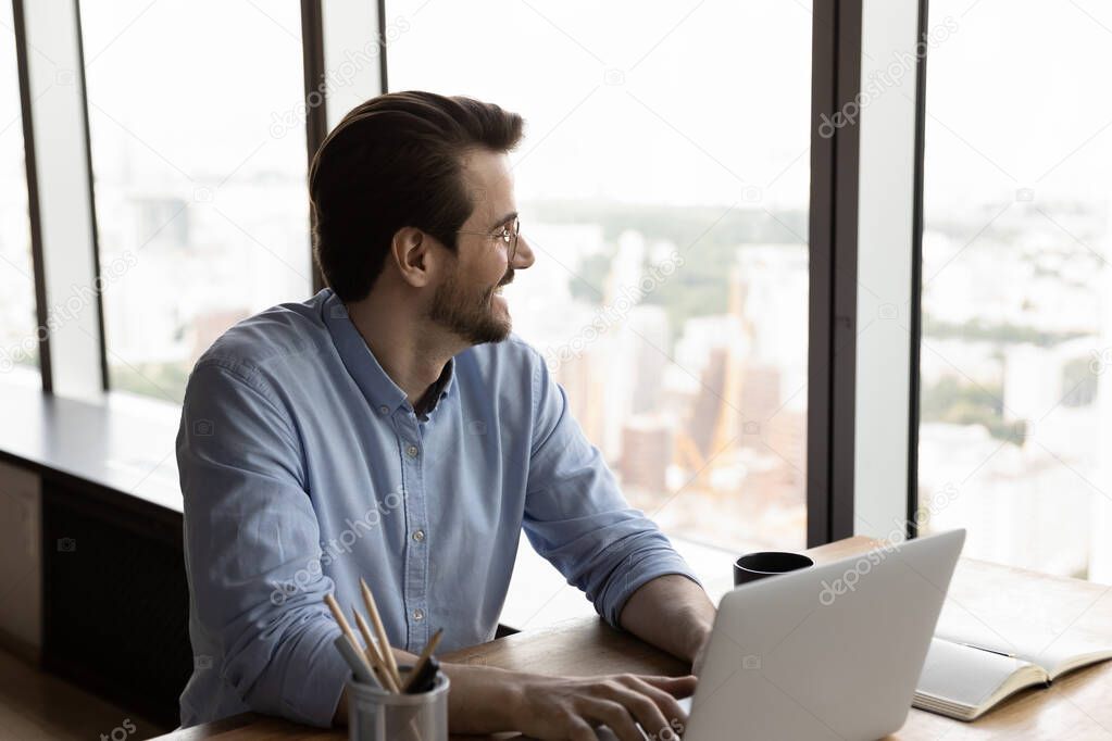 Happy confident thoughtful businessman using laptop, looking out office window