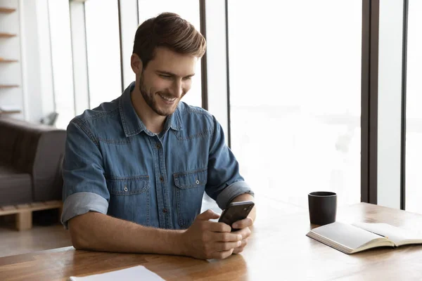 Buon giovane uomo millenario con smartphone seduto a tavola — Foto Stock