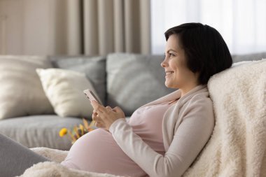 Mutlu genç anne cep telefonu kullanıyor, koltukta dinleniyor.