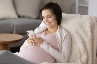 Happy young pregnant woman enjoying leisure at home, using smartphone