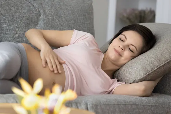 Peaceful sleepy expecting mom resting on couch, caressing bare belly – stockfoto