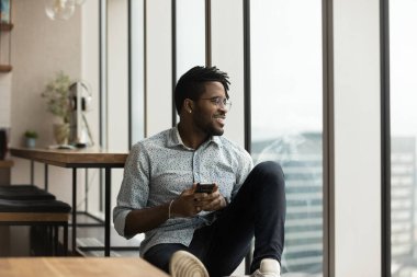 African guy holding smartphone looks at big city through window