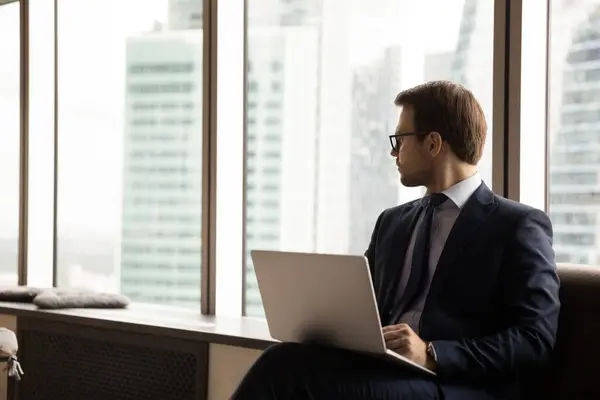 Umsichtiger Geschäftsmann mit Laptop sitzt im Büro und sieht aus wie Wolkenkratzer — Stockfoto
