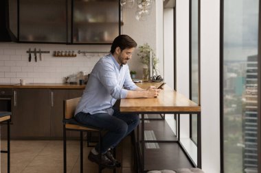 Millennial male sit in modern kitchen using smartphone