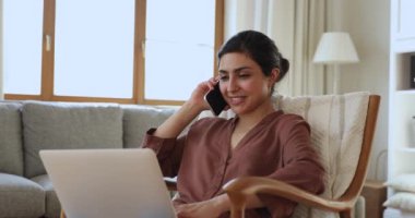 Indian woman talks on smartphone sitting on armchair with laptop