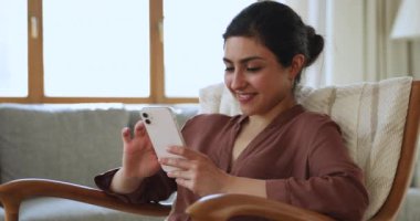 Relaxed Indian woman resting on armchair with smartphone