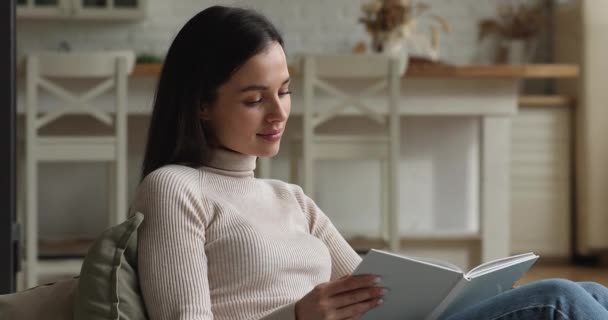 Χαλαρωμένη χαρούμενη νεαρή γυναίκα που διαβάζει χάρτινο βιβλίο στο σπίτι. — Αρχείο Βίντεο