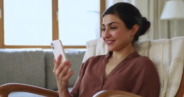 Happy Indian woman relaxing on armchair holding cellphone start videocall
