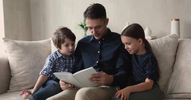 Glücklicher junger Vater liest kleinen Kindern Papierbuch vor. — Stockvideo