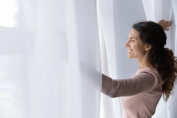 Wanita muda Latina membuka tirai tampak keluar jendela terasa bahagia — Stok Foto